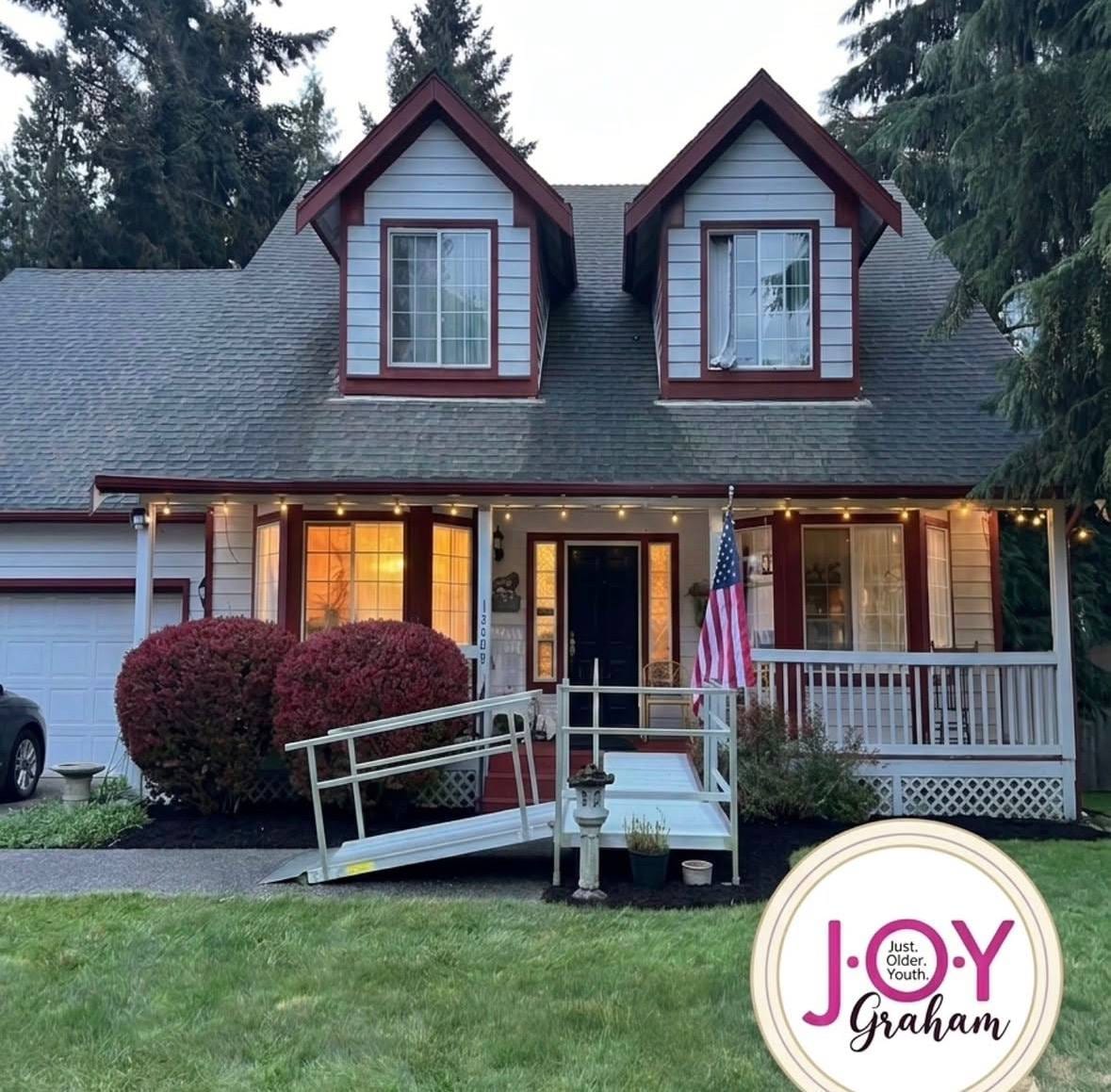 The JOY-Graham home at dusk — welcoming front porch with string lights, American flag, and accessibility ramp.